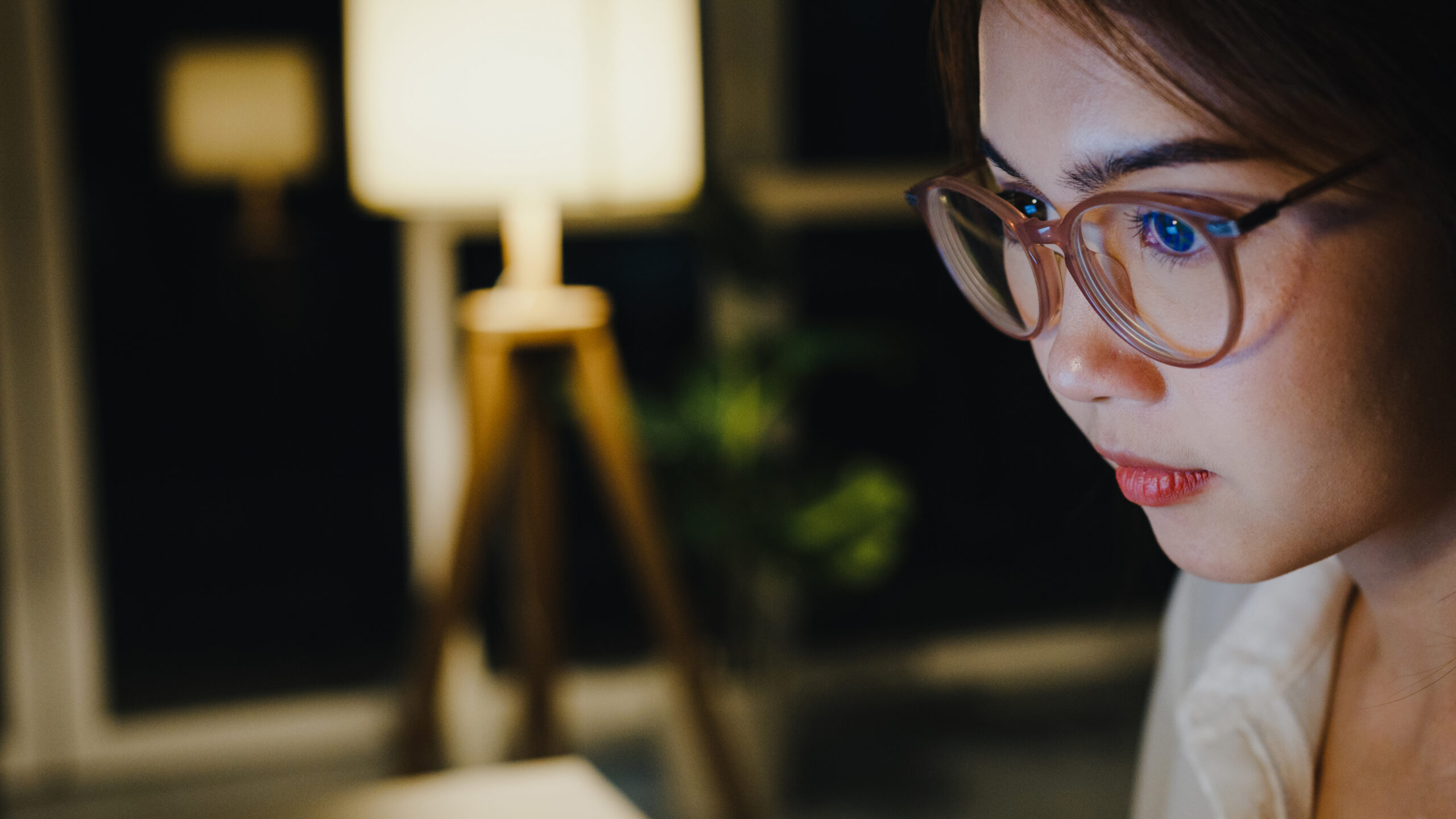 A woman wearing glasses works intently on her laptop at night, illuminated by warm indoor lighting.