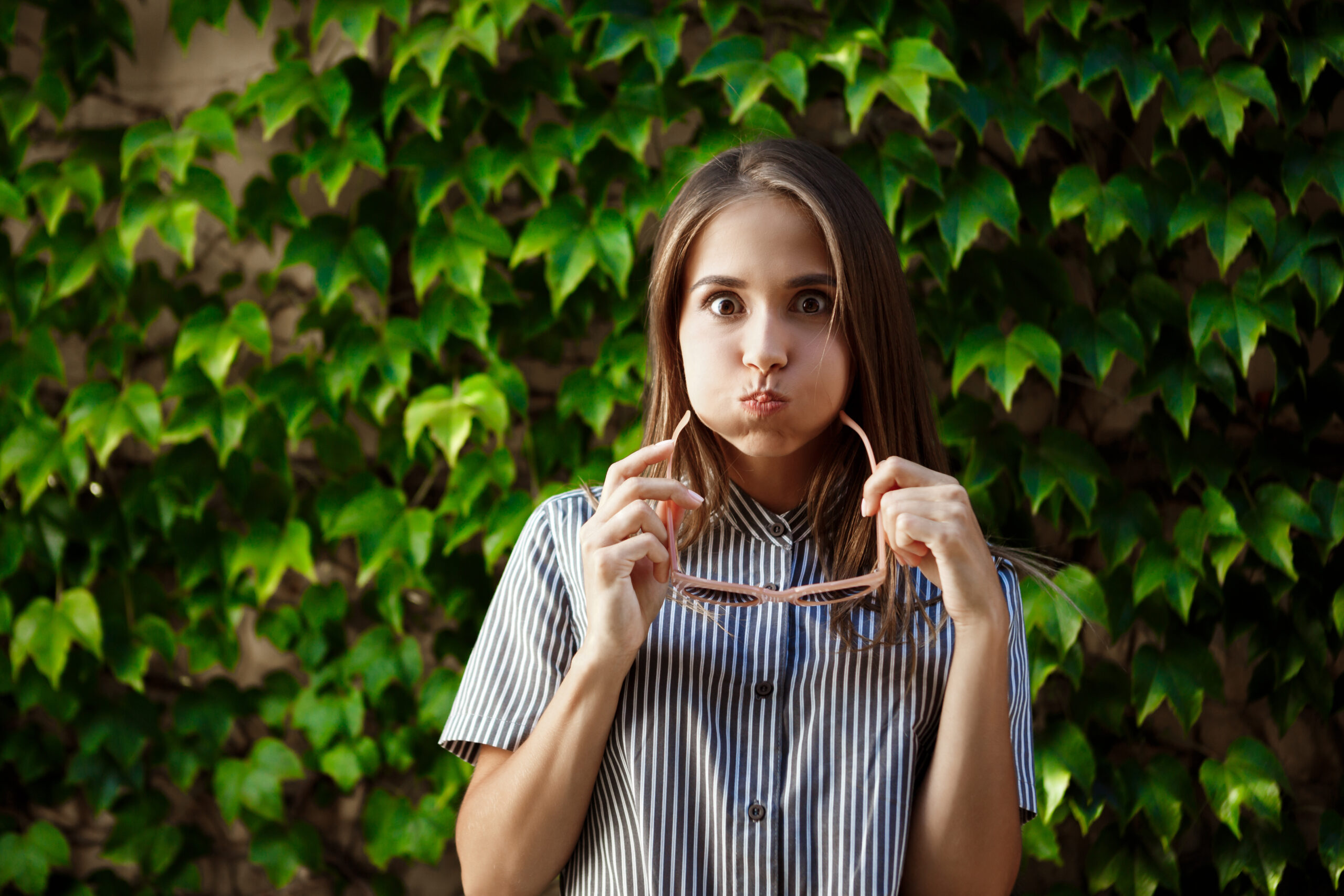 Young woman holding eco-friendly sunglasses while standing in front of a lush green leafy wall, symbolizing sustainability and natural living.