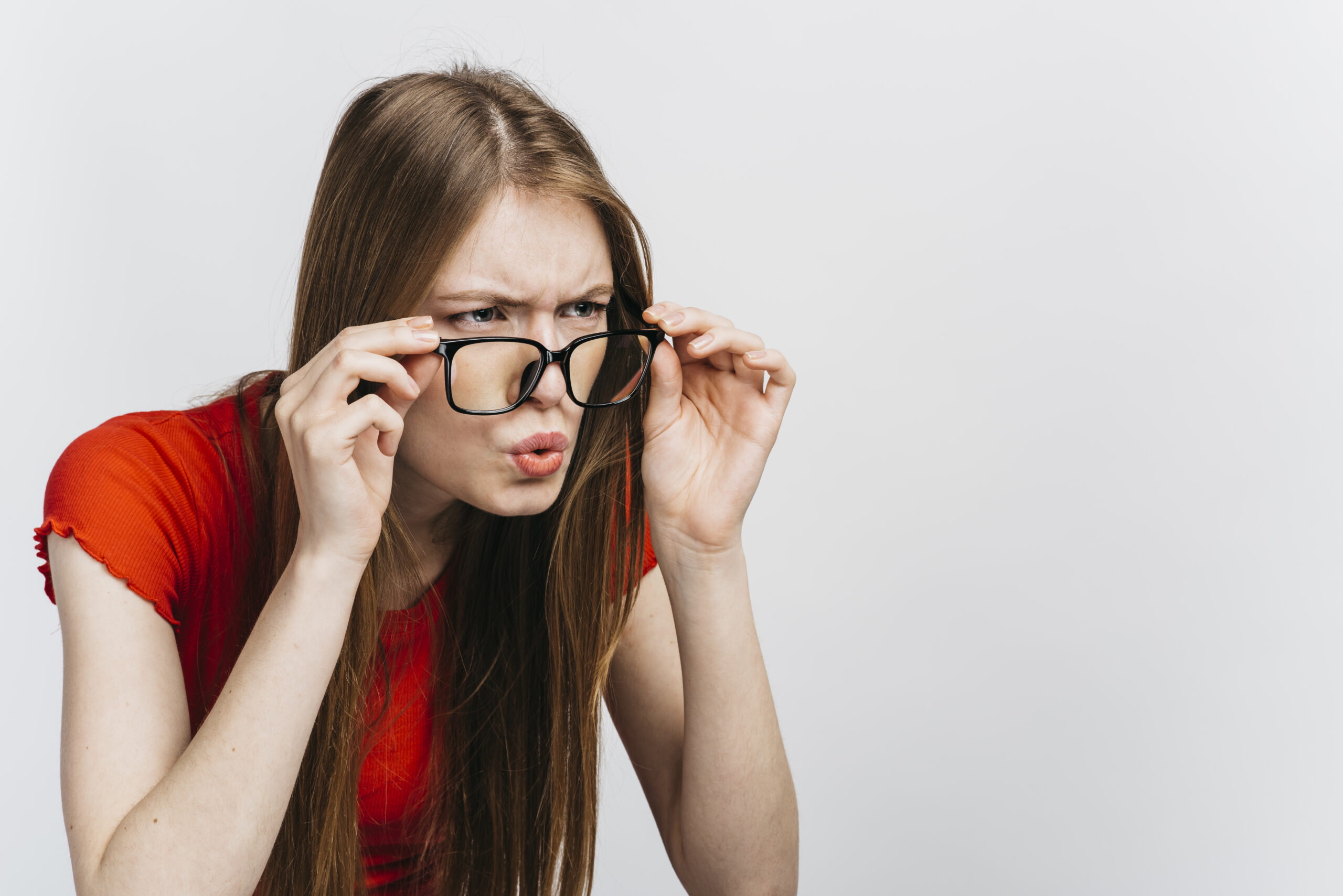 A young woman with long brown hair wearing a red shirt looks intently through her black-framed glasses, leaning forward with a puzzled expression against a plain light background.