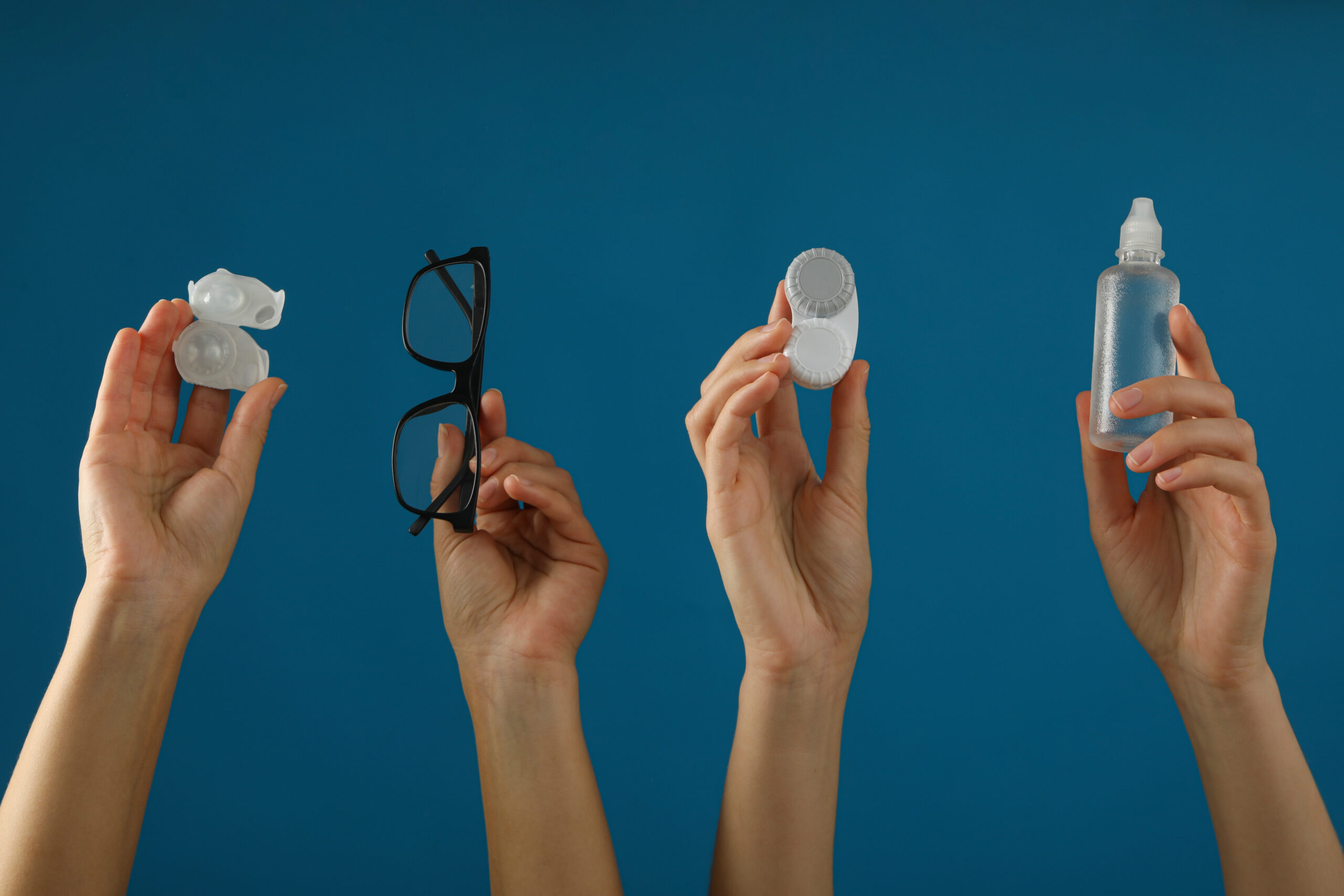 Hands holding eyeglasses, contact lens cases, and a lens solution bottle against a blue background.